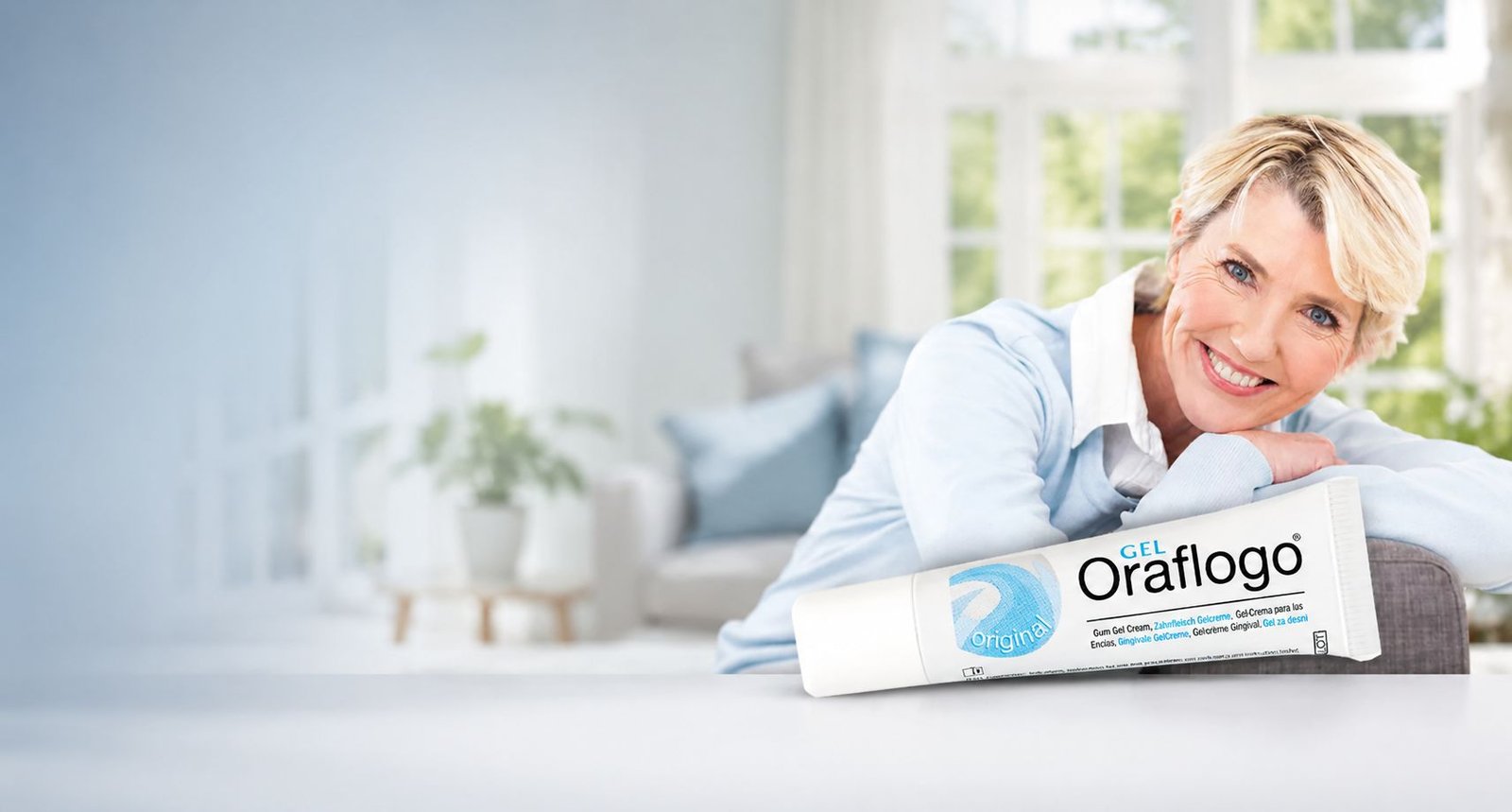 A smiling woman resting beside a tube of Oraflogo Gel in a bright living room